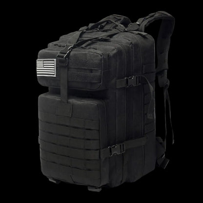 A black tactical military backpack with multiple zippered compartments, MOLLE webbing, compression straps, and a black-and-white American flag patch on the side, shown on a white background.